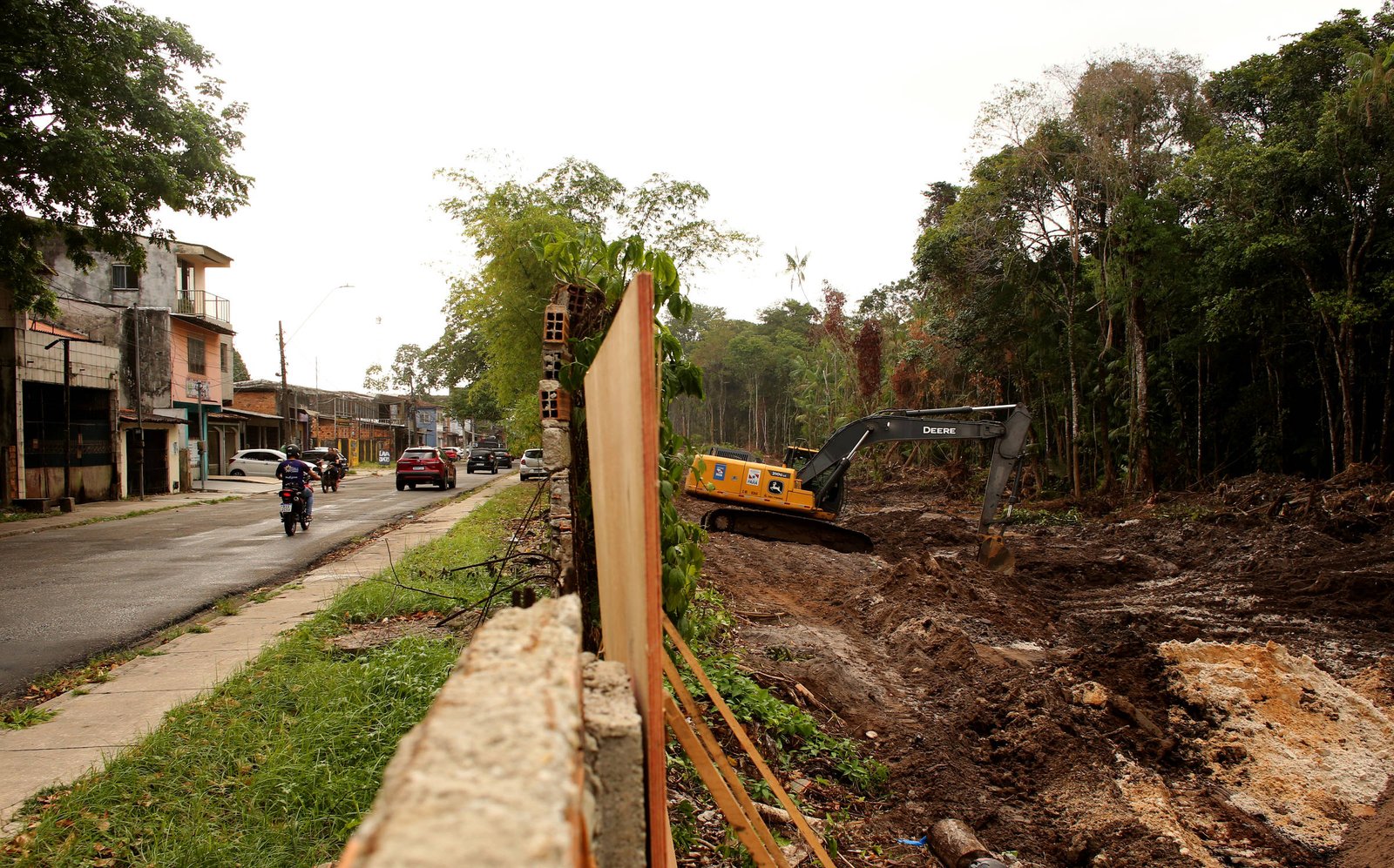 A imagem mostra um canteiro de obras em uma área urbana. À esquerda, há uma rua com uma motocicleta e um carro passando. À direita, o terreno está desmatelado e há uma máquina de construção, possivelmente uma escavadeira, trabalhando. Ao fundo, vê-se uma construção e árvores ao redor.
