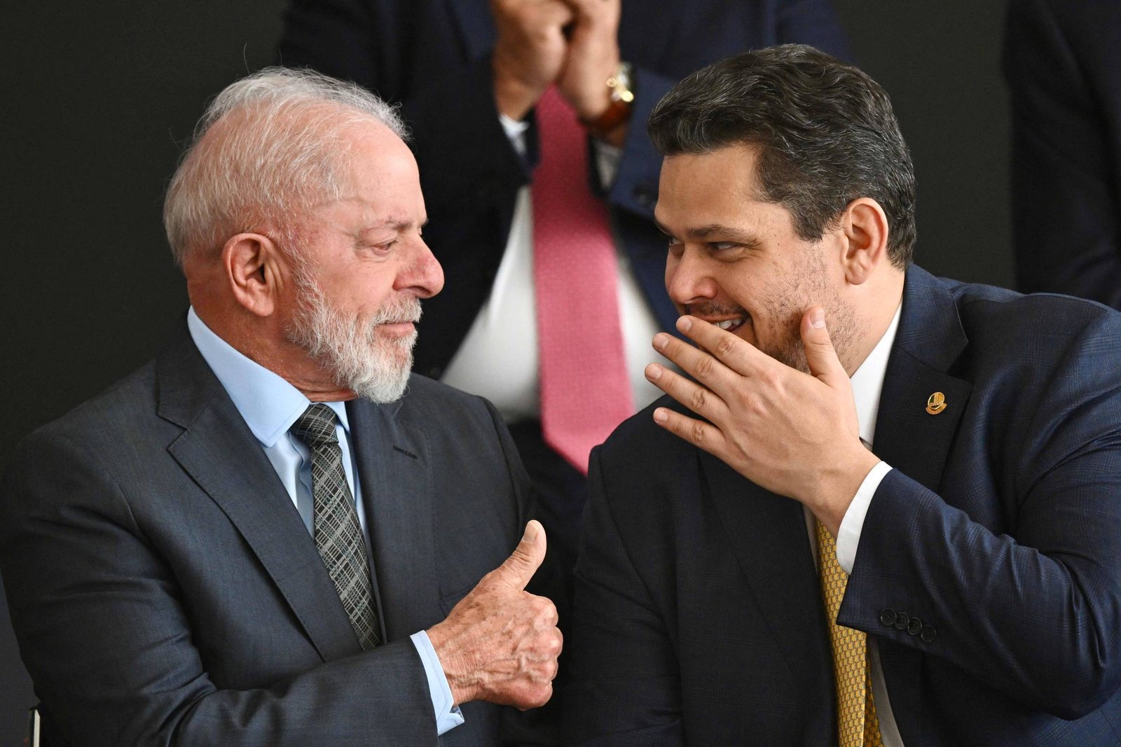 Na imagem, dois homens estão sentados um ao lado do outro, sorrindo e conversando. O homem à esquerda tem cabelo grisalho e usa um terno escuro com uma gravata. O homem à direita tem cabelo escuro e usa um terno claro com uma gravata amarela. Ao fundo, outras pessoas estão parcialmente visíveis, algumas aplaudindo.