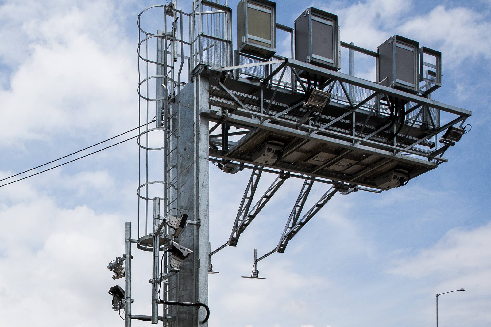 A imagem mostra uma estrutura metálica de sinalização viária, com quatro painéis retangulares na parte superior, possivelmente utilizados para exibir informações de trânsito. A estrutura é apoiada em um poste vertical e possui escada lateral. O céu ao fundo está parcialmente nublado