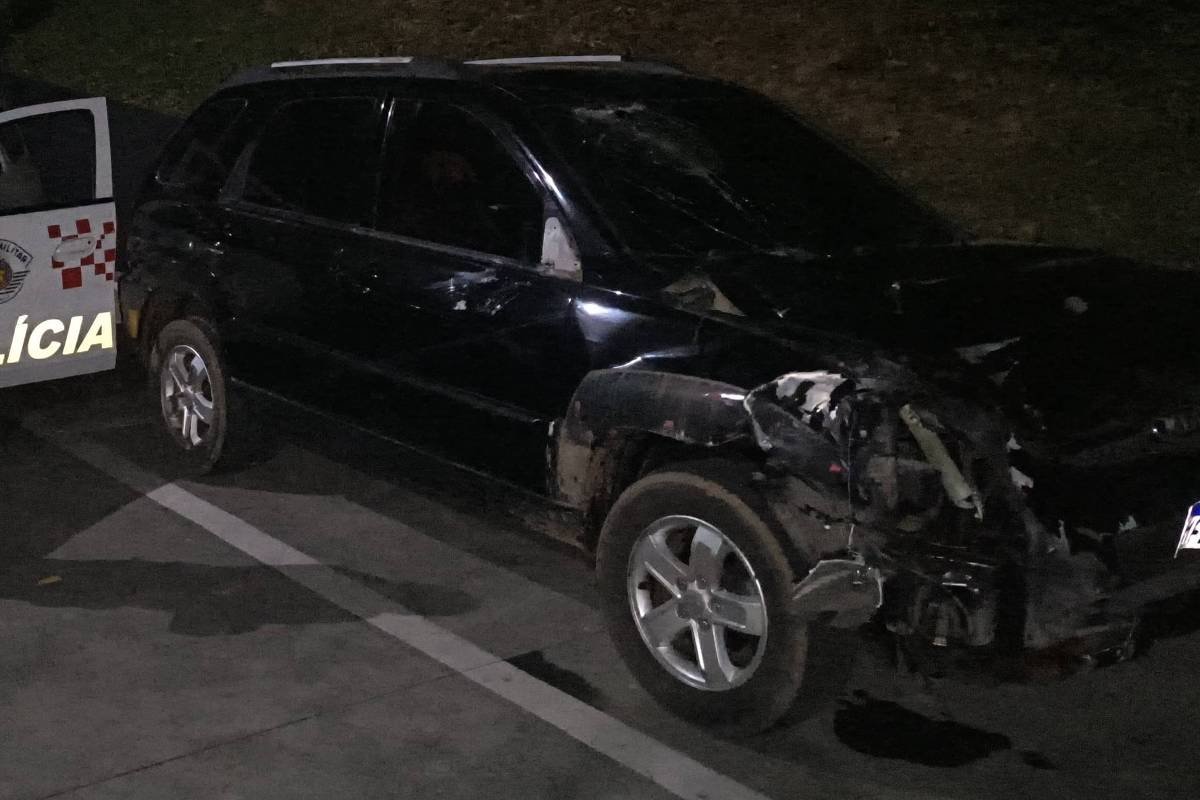 A imagem mostra um policial em pé ao lado de um carro de polícia branco com detalhes em vermelho e azul. O policial está de braços cruzados, usando um uniforme. Ao lado do carro de polícia, há um veículo preto que apresenta danos visíveis na parte frontal. A cena é noturna, com pouca iluminação ao fundo.
