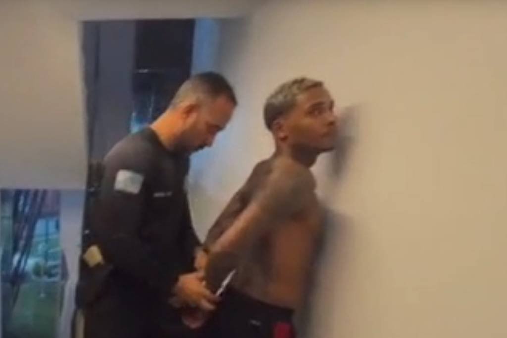 A imagem mostra um homem sem camisa sendo algemado por um policial em um ambiente interno. O homem está encostado na parede, enquanto outro policial observa. Há roupas no chão e uma janela ao fundo, sugerindo um ambiente residencial.