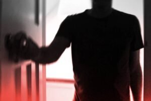 A imagem mostra uma silhueta de uma pessoa com um braço estendido, segurando uma maçaneta de porta. O fundo é desfocado e apresenta uma iluminação suave, com um efeito de gradiente que vai do vermelho na parte inferior ao preto e branco na parte superior. A figura da pessoa é parcialmente visível, destacando-se contra o fundo.