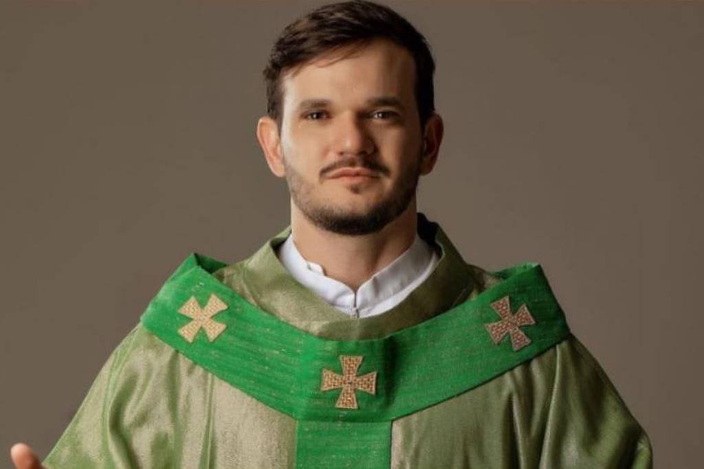 Um homem está vestido com uma túnica litúrgica verde, adornada com cruzes douradas. Ele está em pé, com as mãos levantadas em um gesto de bênção ou saudação. O fundo é neutro, destacando a figura do homem.
