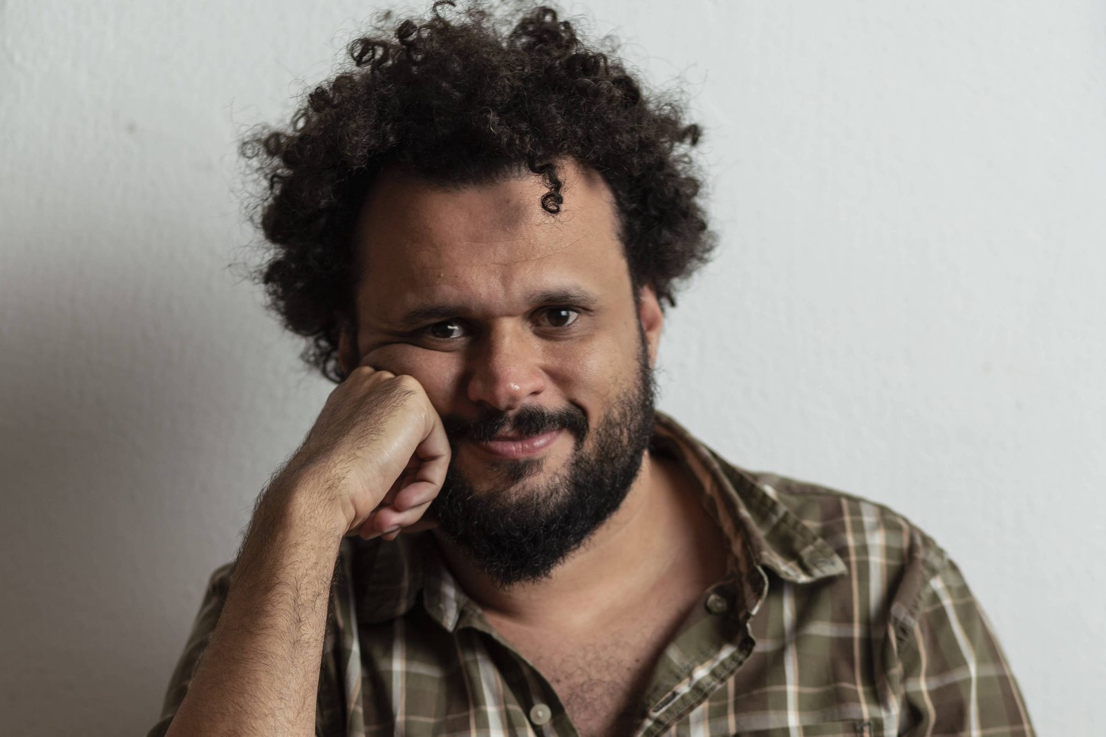 Um homem com cabelo cacheado e barba, sentado à mesa, apoiando o queixo na mão. Ele usa uma camisa xadrez em tons de verde e marrom. O fundo é uma parede branca, e a mesa é de madeira.