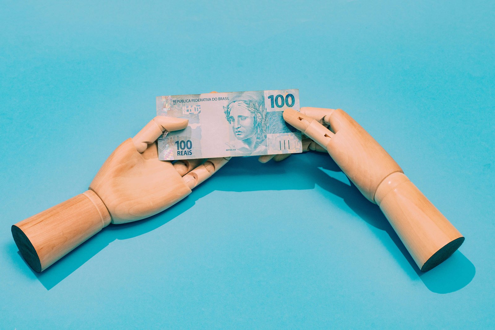 A imagem mostra duas mãos de madeira, uma de cada lado, segurando uma nota de 100 reais. O fundo é azul, destacando as mãos e a nota. A nota apresenta a figura de uma mulher e o valor de 100 em destaque.