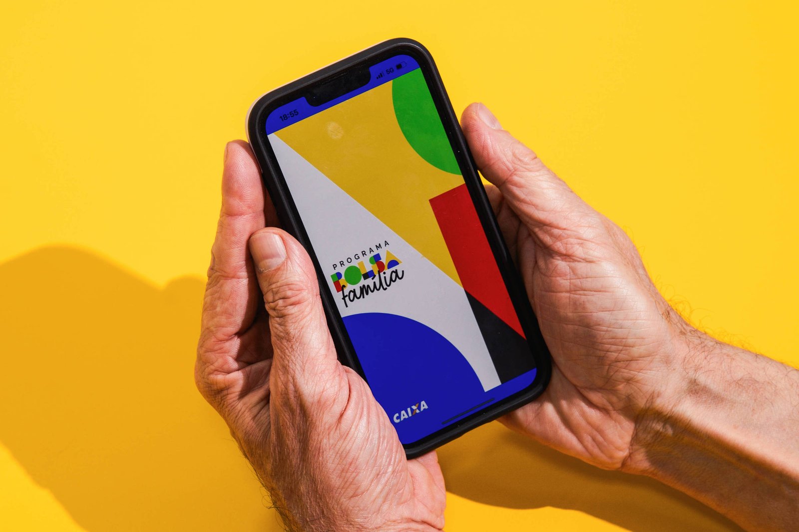 A imagem mostra um smartphone sendo segurado por duas mãos. A tela do smartphone exibe um design colorido com formas geométricas em azul, verde, amarelo e vermelho. O fundo da imagem é amarelo, criando um contraste vibrante com o dispositivo.