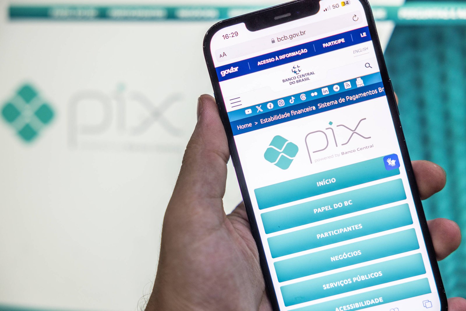 A imagem mostra um smartphone sendo segurado por uma mão, exibindo a tela do aplicativo Pix. O fundo apresenta o logotipo do Pix em uma superfície clara. A tela do smartphone contém opções relacionadas ao serviço de pagamento, com botões em azul e branco.