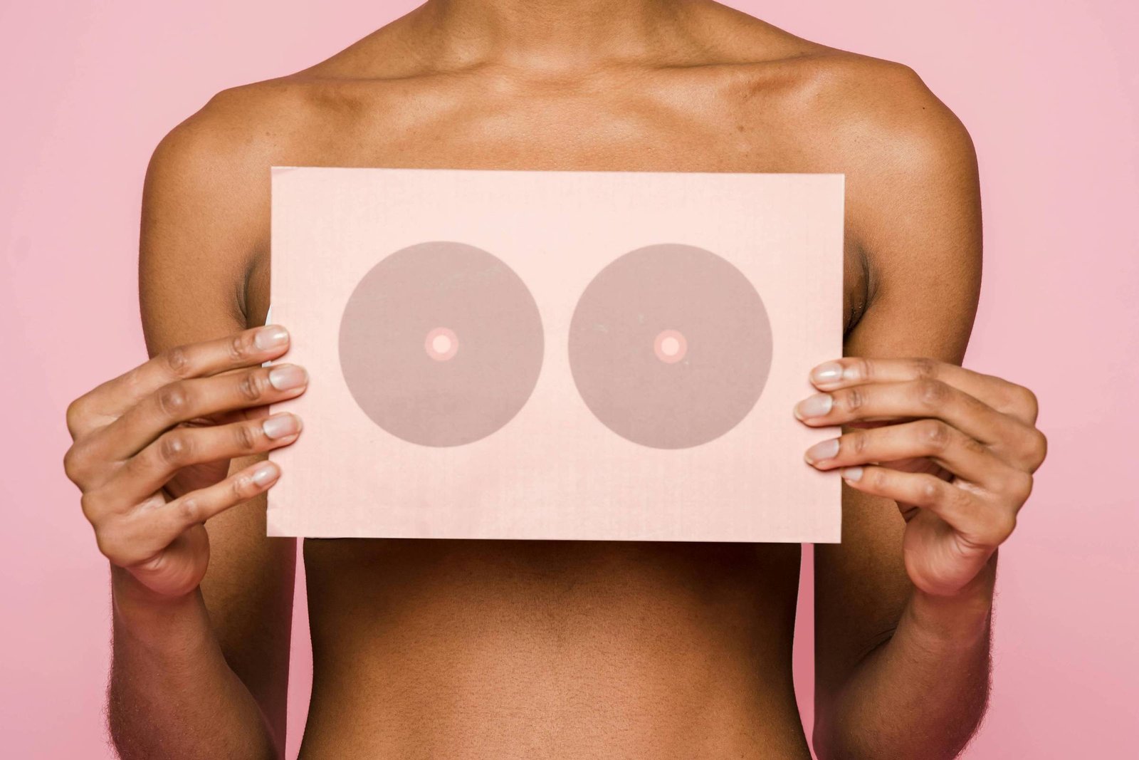 Uma mulher sorridente está segurando um cartaz que possui dois círculos que representam seios. Ela está sem blusa, com a pele exposta, e o fundo é de cor rosa. A mulher tem cabelo curto e está em uma pose descontraída.