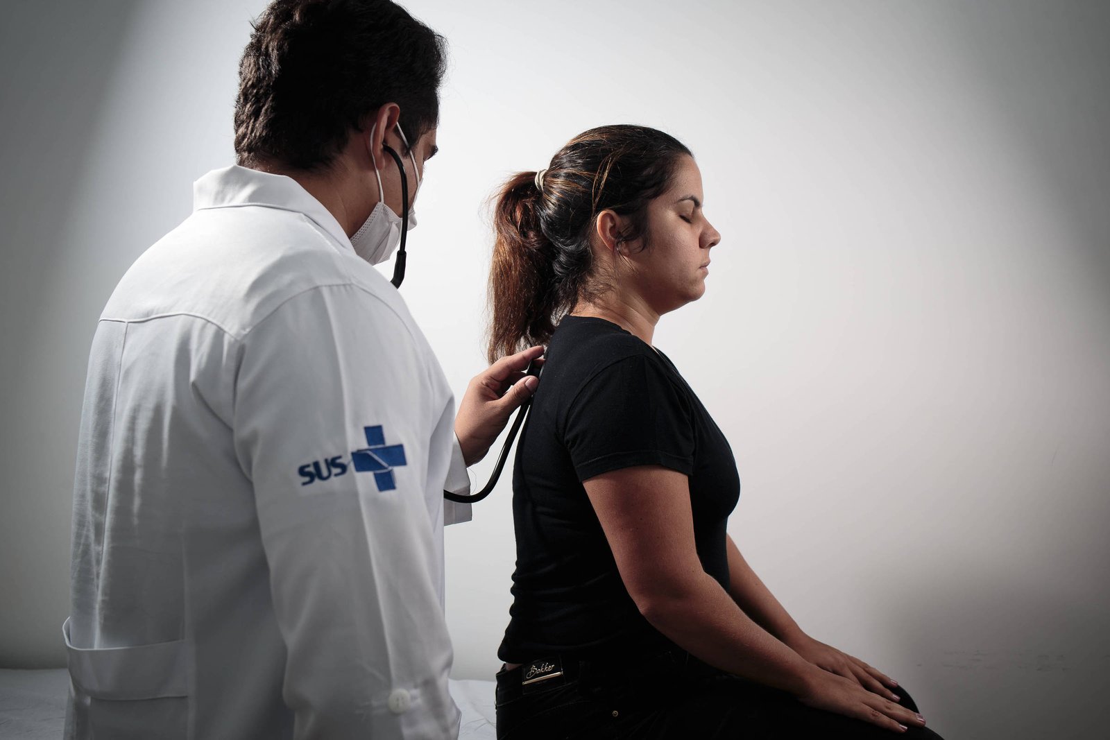 A imagem mostra um médico, vestido com um jaleco branco e usando uma máscara, auscultando uma paciente que está sentada. A paciente é uma mulher com cabelo preso, vestindo uma camiseta preta. O ambiente é neutro, com fundo claro.
