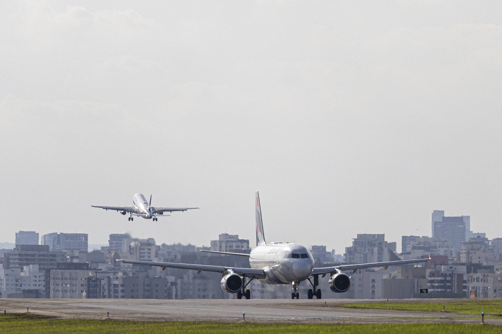 A imagem mostra dois aviões em um aeroporto. Um avião está taxiando na pista, enquanto outro está pousando ao fundo, com a cidade visível ao longe. O céu está parcialmente nublado e a área ao redor é urbana, com prédios e estruturas visíveis.