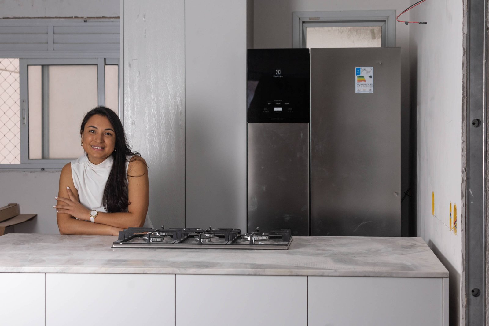 Uma mulher está sentada em uma bancada de cozinha moderna, com um fogão a gás na frente. Ao fundo, há uma geladeira prateada e uma janela com grades. A mulher tem cabelo longo e liso, e usa uma blusa branca. O ambiente é bem iluminado e possui paredes claras.