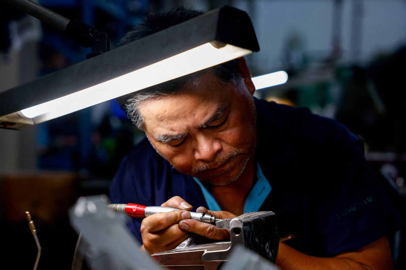 Um homem está concentrado em seu trabalho em uma oficina, utilizando uma ferramenta para realizar um ajuste em uma peça metálica. Ele está sob uma luz intensa que ilumina sua área de trabalho, e seu rosto demonstra foco e dedicação. O ambiente ao fundo é desfocado, mas sugere um espaço de trabalho industrial.