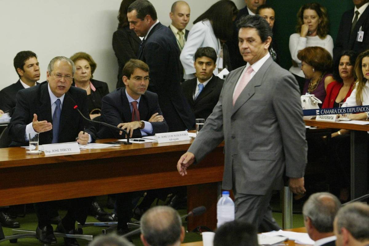 A imagem mostra uma audiência na Câmara dos Deputados do Brasil. Vários homens e mulheres estão sentados em mesas, alguns com expressões sérias, enquanto um homem de terno claro caminha em direção ao centro da sala. O ambiente é formal, com uma mesa longa à frente e pessoas observando ao fundo. Há também um painel com a inscrição