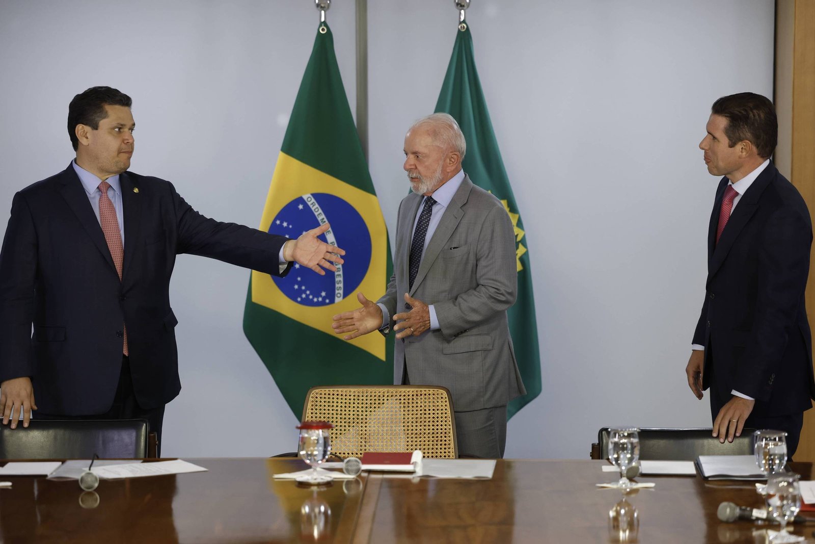 A imagem mostra três homens em uma mesa de reunião. Dois deles estão de pé, um à esquerda, vestido com um terno escuro e outro à direita, também de terno, com uma gravata vermelha. O homem no meio, que está de mãos levantadas, usa um terno claro. Ao fundo, estão as bandeiras do Brasil. A mesa está decorada com copos de vidro e um caderno.