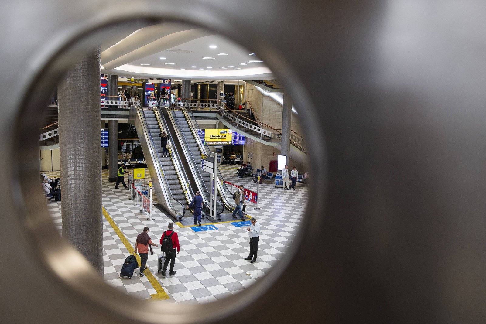 A imagem mostra a vista de um terminal de aeroporto através de um buraco circular. No interior, há escadas rolantes e várias pessoas se movendo pelo espaço. O piso é de ladrilhos em padrão quadriculado, e há sinalizações visíveis, incluindo anúncios. O ambiente parece bem iluminado e moderno.