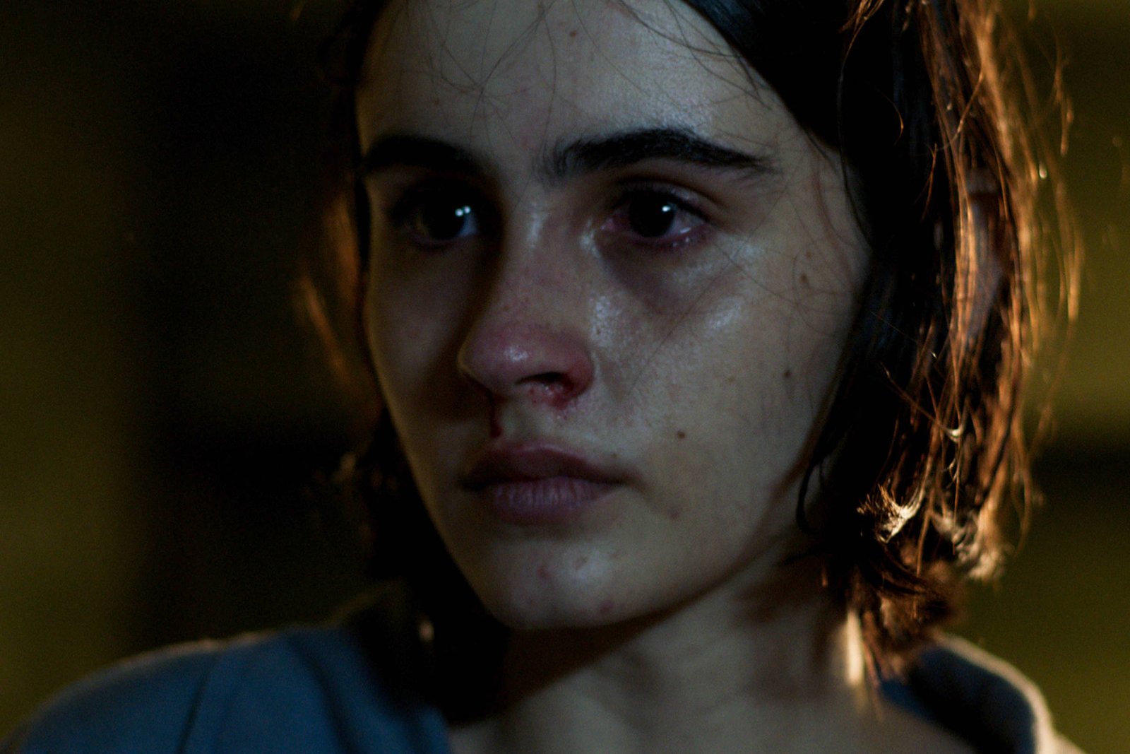 A imagem mostra uma jovem com cabelo curto e escuro, com uma expressão de preocupação ou tristeza. Ela tem lágrimas nos olhos e a pele levemente brilhante, sugerindo que está emocionada. O fundo é desfocado, com pouca iluminação, o que destaca seu rosto.