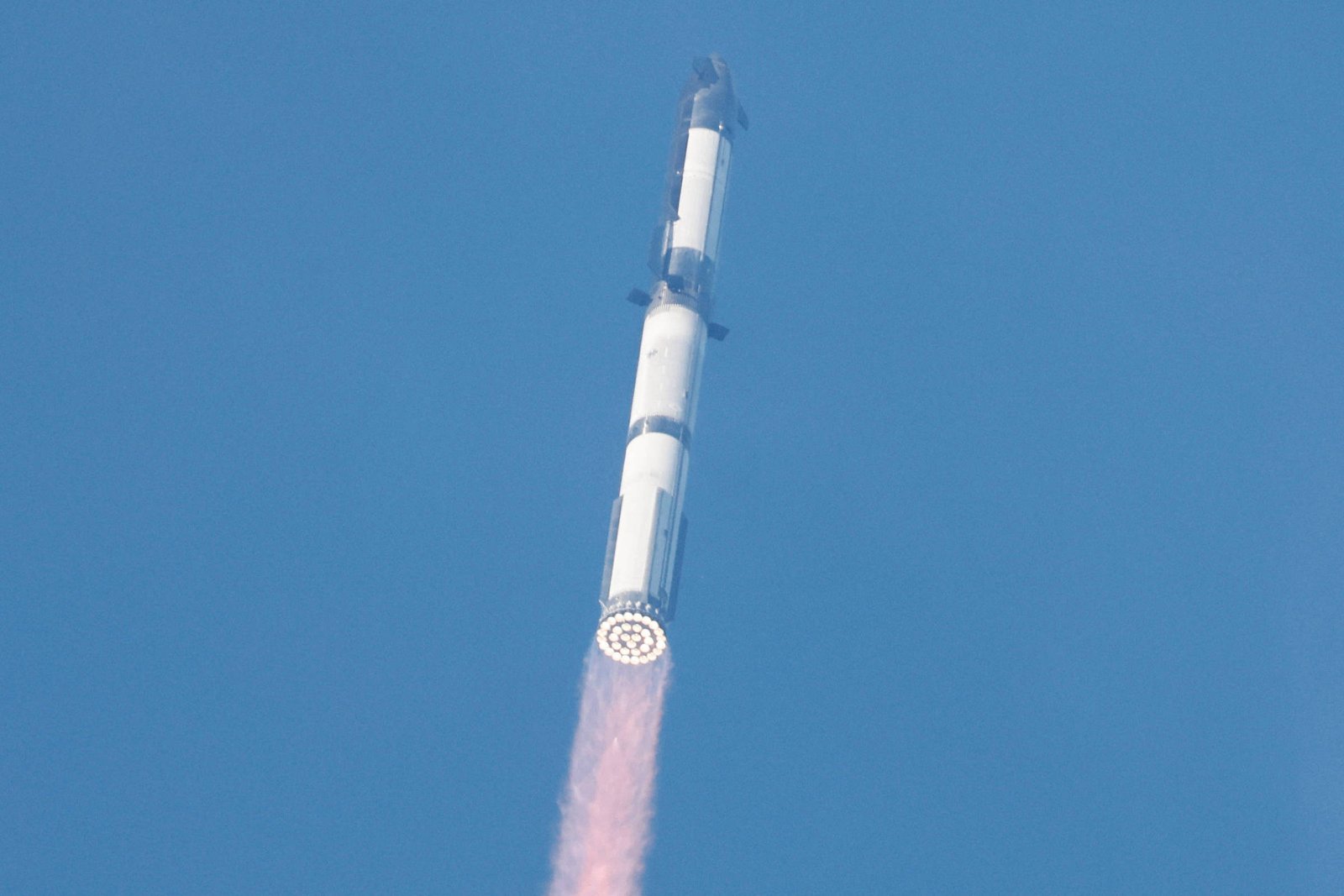 A imagem mostra um foguete subindo em direção ao céu, com uma trilha de fumaça rosa saindo de sua base. O fundo é azul, indicando um dia claro.
