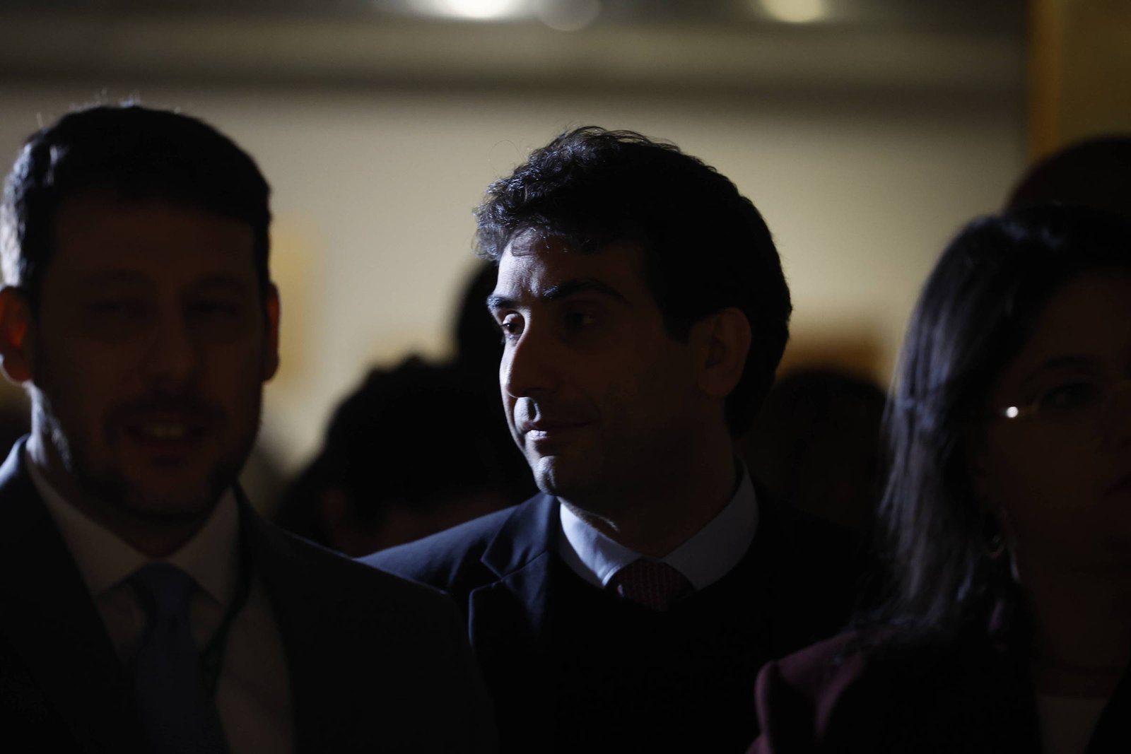 A imagem mostra um grupo de pessoas em um ambiente com pouca luz. Um homem em destaque, com cabelo escuro e liso, está olhando para o lado. Ele usa um paletó escuro e uma camisa clara. À sua esquerda, um homem sorridente com cabelo curto e barba está parcialmente visível. À direita, uma mulher com cabelo solto e óculos também está presente, mas seu rosto não está totalmente iluminado. O fundo é desfocado, sugerindo um ambiente de evento ou reunião.