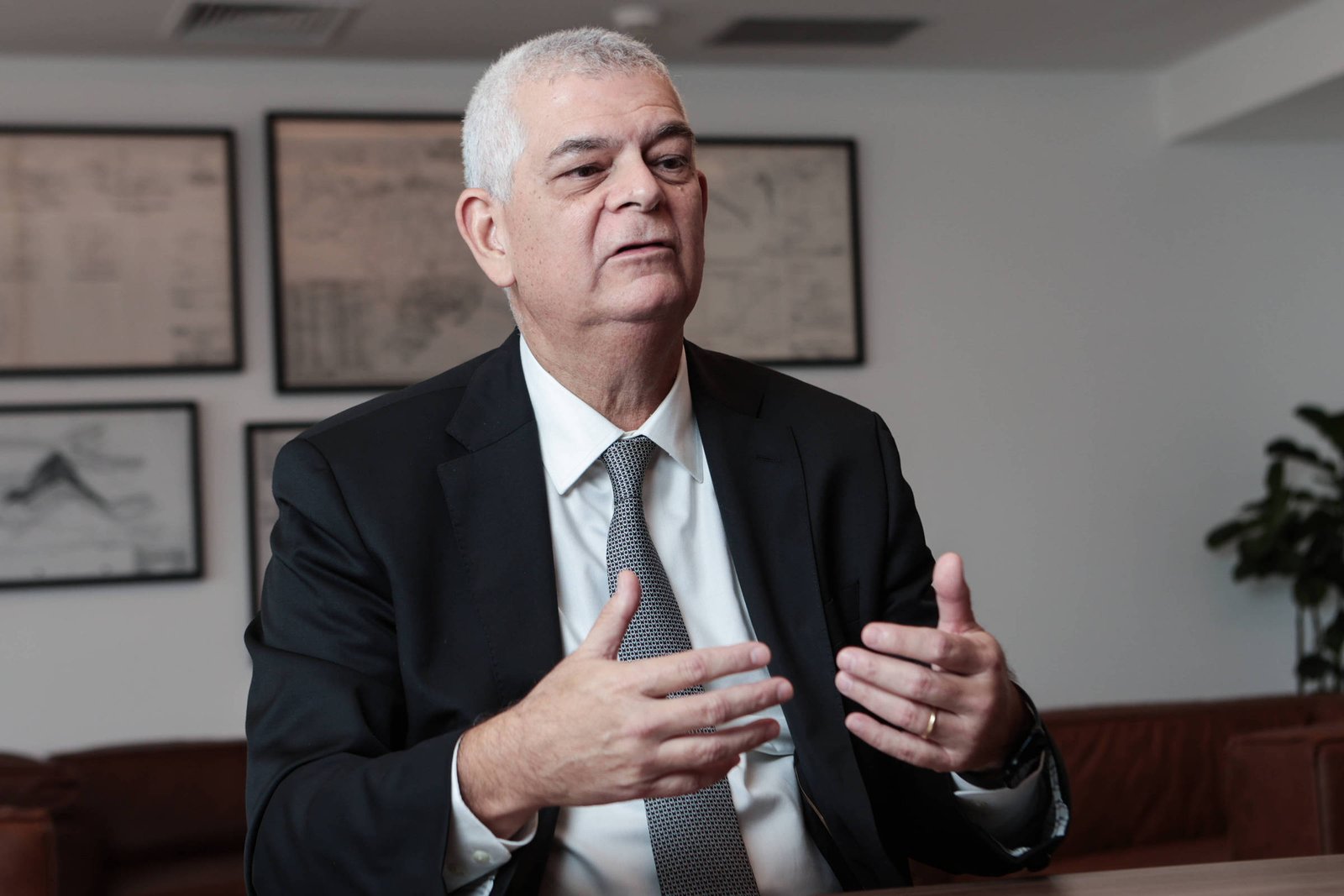 Um homem de cabelos grisalhos, vestido com um terno preto e uma gravata prateada, está sentado em uma mesa de escritório. Ele está gesticulando com as mãos enquanto fala. Ao fundo, há quadros com desenhos ou gráficos nas paredes e uma planta em um canto. O ambiente parece moderno e bem iluminado.