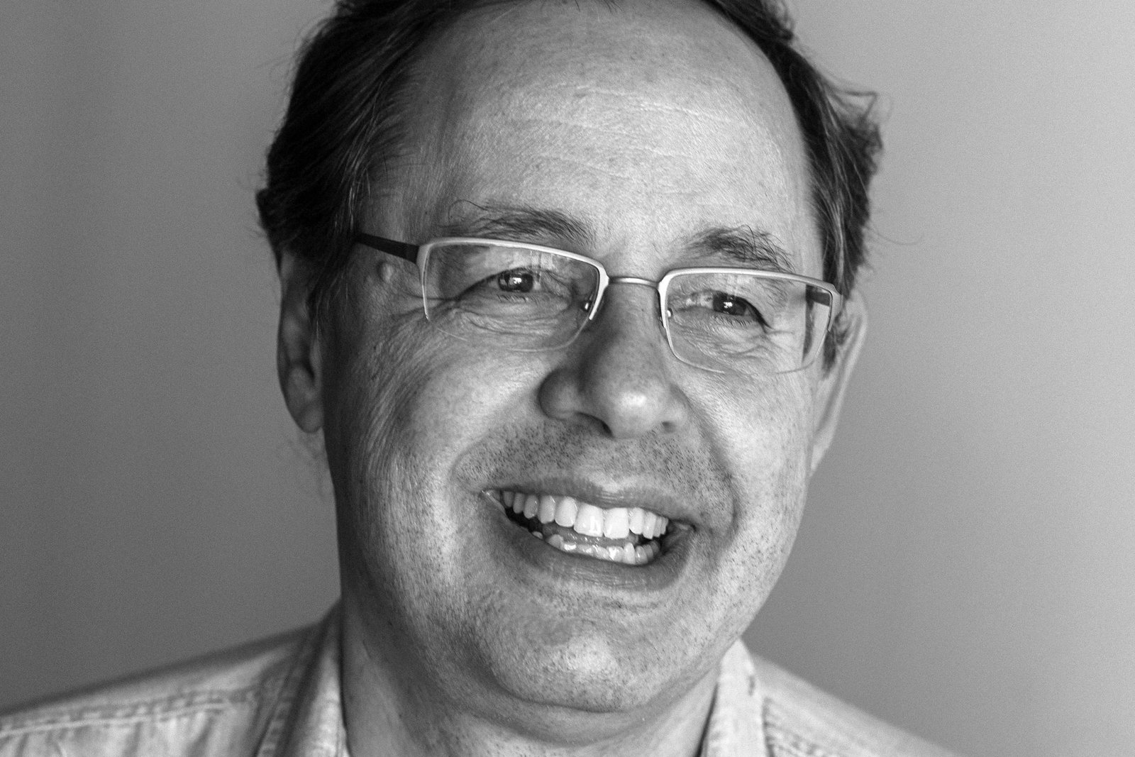 A imagem mostra um homem sorridente em um retrato em preto e branco. Ele está usando uma camisa clara com pequenos detalhes e óculos. O fundo é neutro e desfocado, destacando o rosto do homem.