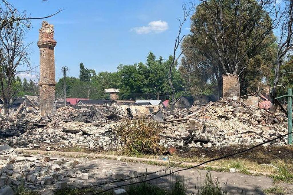 A imagem mostra os restos de um edifício em ruínas, com paredes de tijolos e escombros espalhados pelo chão. Dois chaminés de tijolos permanecem em pé, cercadas por árvores parcialmente queimadas. O céu está claro, com algumas nuvens, e ao fundo, é possível ver outras construções e vegetação.