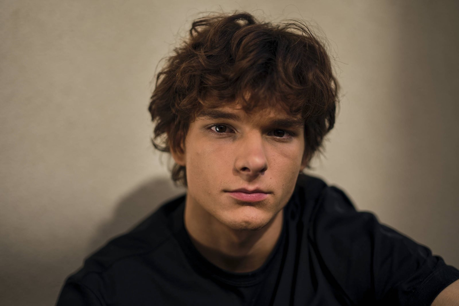 A imagem mostra um jovem com cabelo cacheado e bagunçado, olhando diretamente para a câmera. Ele está vestindo uma camiseta preta e parece estar em um ambiente com iluminação suave, que destaca seu rosto. O fundo é de uma cor neutra, o que faz com que o foco permaneça no jovem.