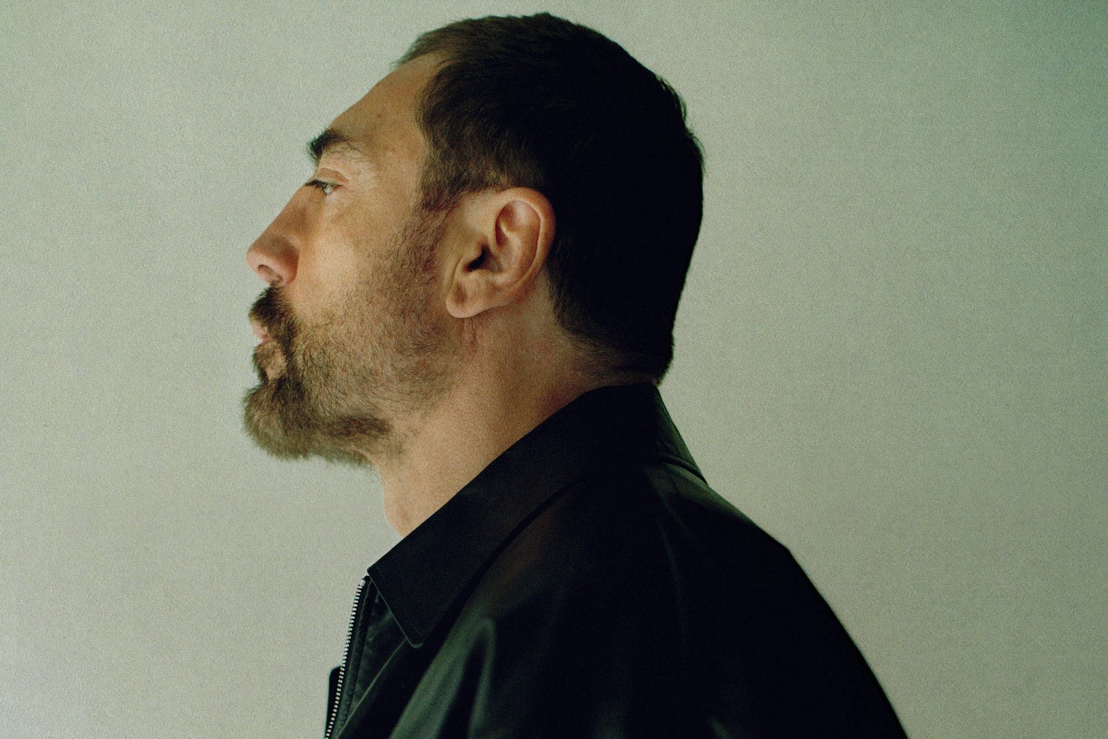 A imagem mostra um homem em perfil, com cabelo escuro e barba. Ele está usando uma jaqueta preta e está posicionado de lado, olhando para a esquerda. O fundo é de uma cor clara e neutra, destacando a figura do homem.