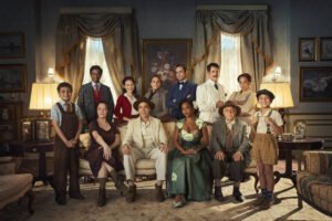 A imagem mostra um grupo de pessoas posando em um ambiente interno elegante, com cortinas e quadros nas paredes. Há um total de 13 pessoas, incluindo crianças e adultos, vestindo roupas de época. Algumas pessoas estão em pé, enquanto outras estão sentadas. O ambiente é bem iluminado, com duas lâmpadas acesas ao fundo.
