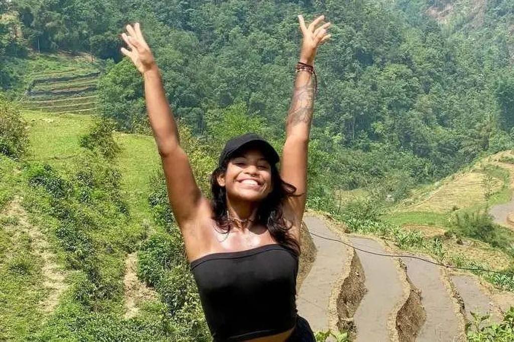 Uma mulher está em uma paisagem montanhosa, com as mãos levantadas em sinal de alegria. Ela usa uma blusa preta e um boné. Ao fundo, há campos de arroz em terraços e vegetação verdejante, com montanhas ao longe.