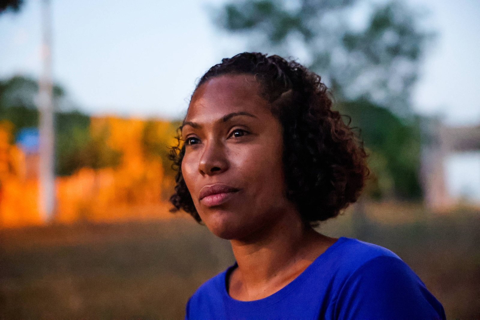 A imagem mostra uma mulher com cabelo cacheado e curto, usando uma blusa azul. Ela está olhando para o lado, com uma expressão pensativa. O fundo apresenta uma paisagem desfocada, com árvores e uma iluminação suave, sugerindo que a foto foi tirada ao entardecer.