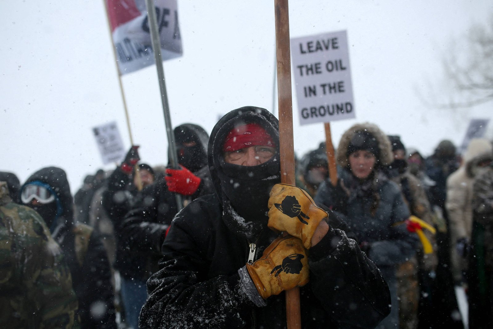 A imagem mostra um grupo de manifestantes em um ambiente nevado. Um homem em primeiro plano segura um bastão e usa luvas amarelas, com o rosto coberto por um cachecol e um gorro. Ao fundo, outros manifestantes também estão presentes, alguns segurando cartazes. Um dos cartazes é visível e diz
