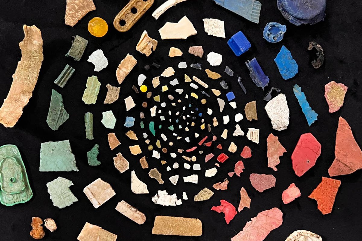 A imagem apresenta uma disposição circular de diversos fragmentos de materiais, incluindo pedaços de cerâmica, vidro e outros materiais coloridos. Os fragmentos variam em formas e tamanhos, com cores que vão do verde, azul, vermelho, amarelo, branco e preto. O fundo da imagem é preto, o que destaca ainda mais as cores vibrantes dos fragmentos dispostos em espiral.
