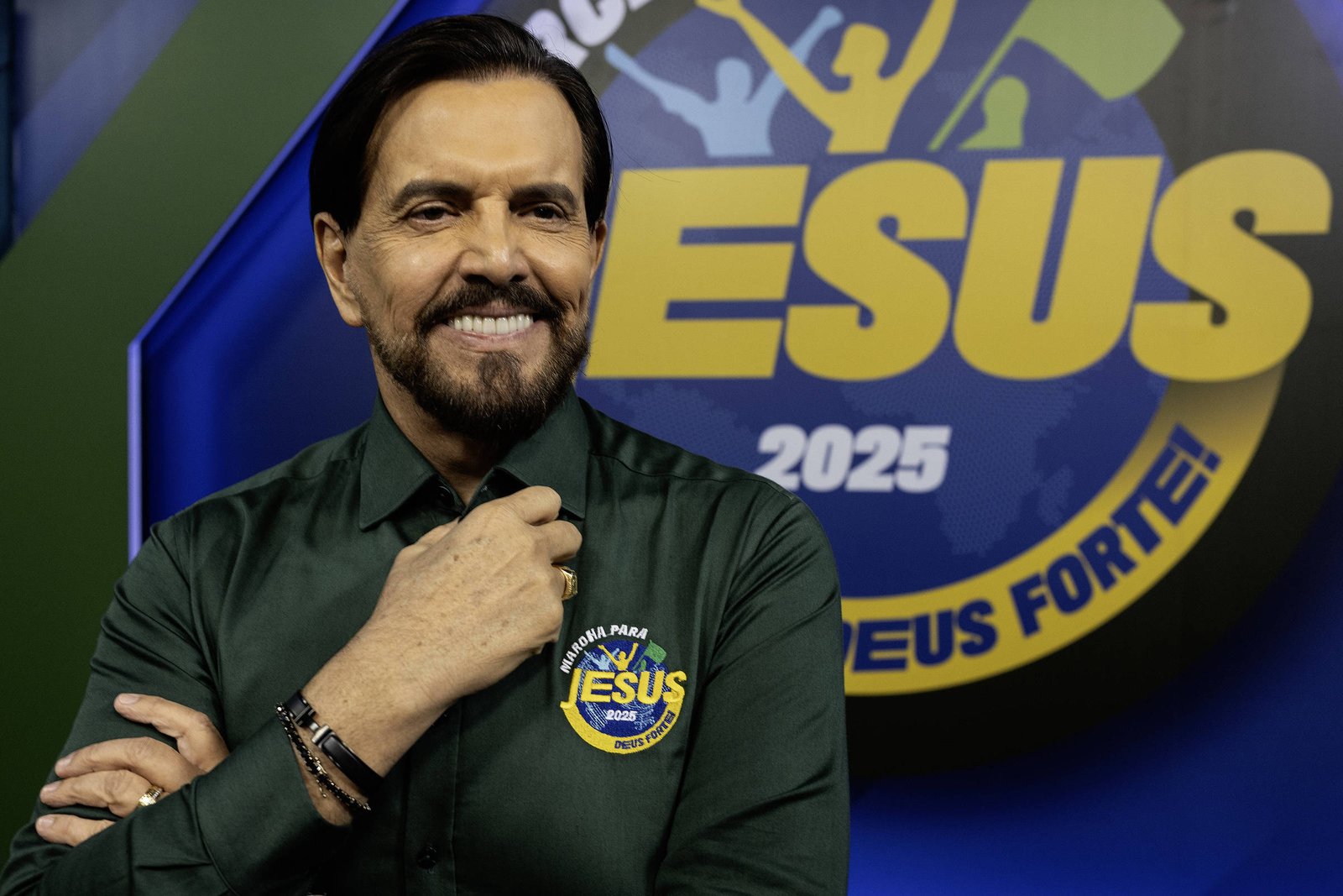Um homem sorridente está posando para a foto em um estúdio. Ele usa uma camisa verde escura e está com a mão no queixo. Ao fundo, há um logotipo colorido que diz