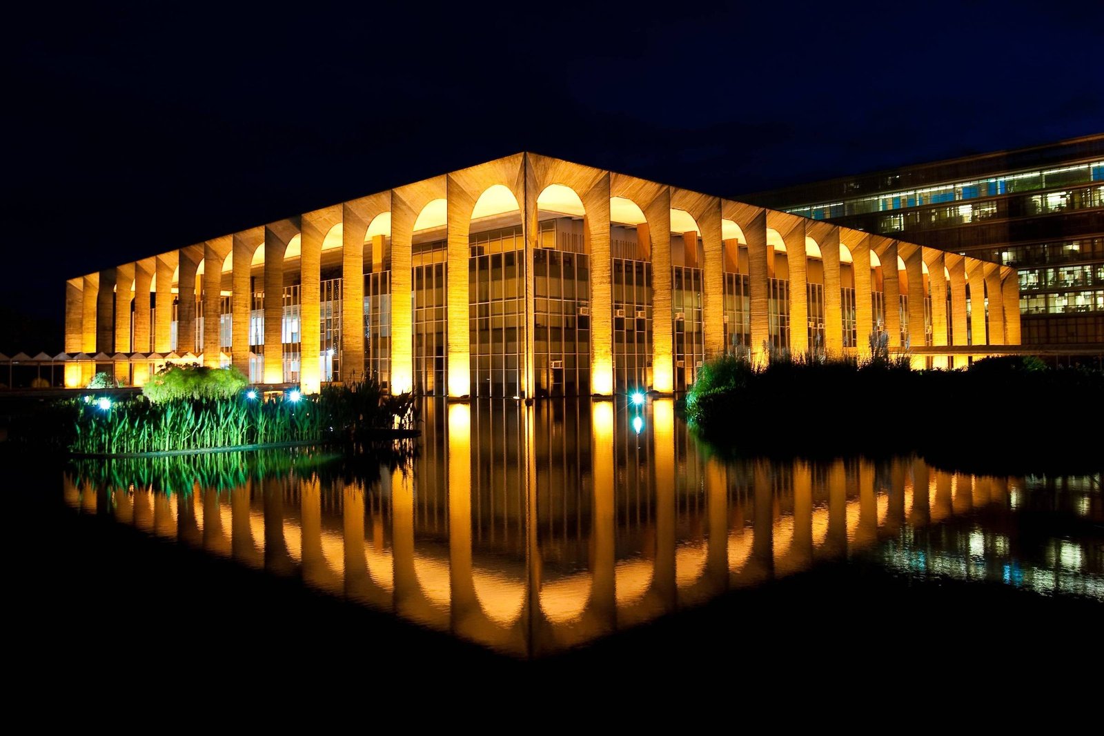 A imagem mostra um edifício moderno iluminado à noite, com uma fachada de colunas e grandes janelas. O edifício reflete na água em frente, criando um efeito visual impressionante. A iluminação destaca a estrutura e a vegetação ao redor, que inclui plantas verdes e um pequeno lago.