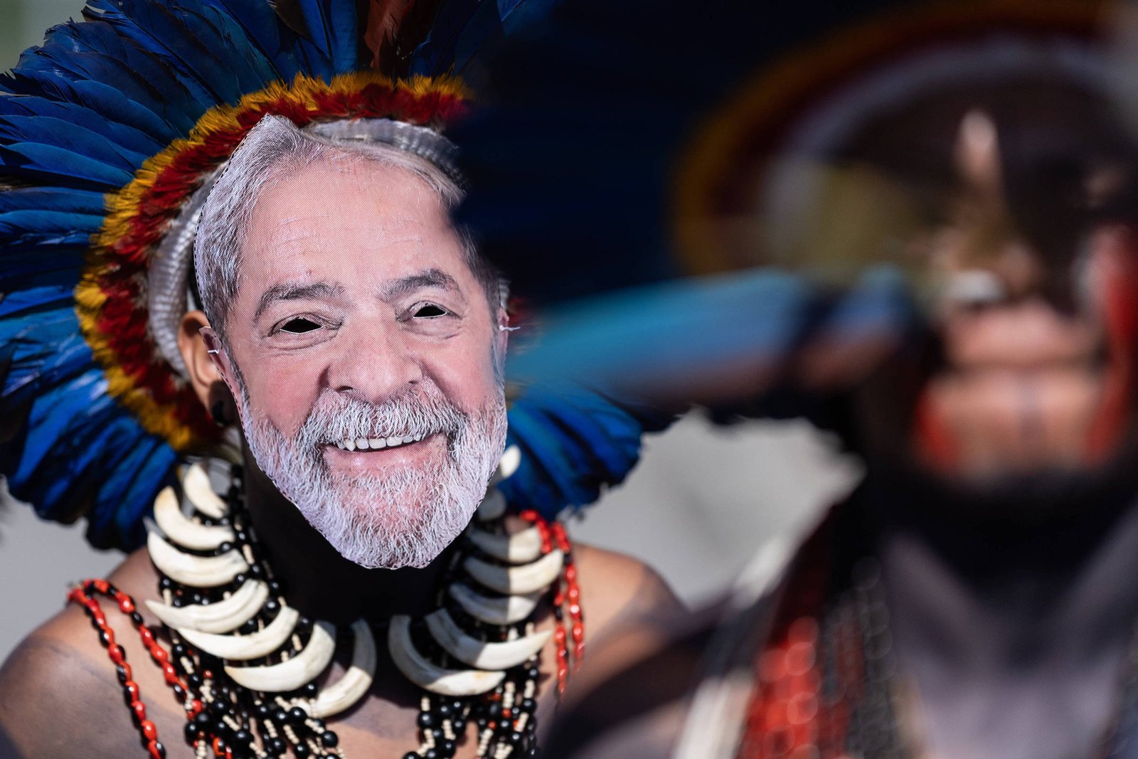 A imagem mostra uma pessoa vestida com trajes indígenas, incluindo um cocar colorido e colares. O rosto da pessoa é coberto por uma máscara que retrata um homem idoso com barba e sorriso, possivelmente uma figura pública. Ao fundo, há outra pessoa com características semelhantes, mas menos visível. A cena é vibrante e destaca a cultura indígena.