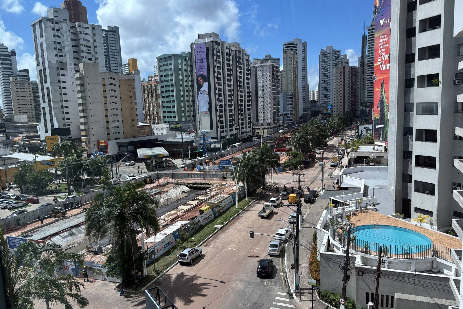 A imagem mostra uma vista panorâmica de uma área urbana com muitos edifícios altos. No primeiro plano, há uma rua com alguns carros e palmeiras. Ao fundo, é possível ver um canteiro de obras e mais edifícios. O céu está claro com algumas nuvens.