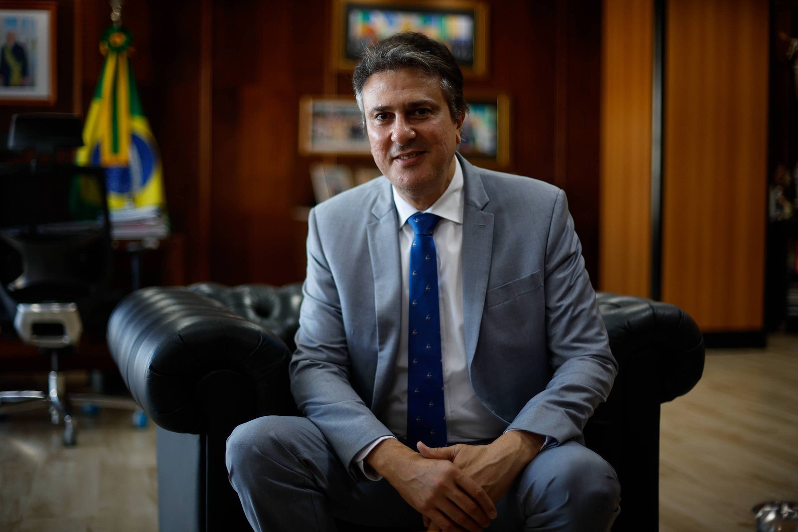 Um homem sentado em um sofá de couro preto em um escritório. Ele está vestindo um terno cinza claro, uma camisa branca e uma gravata azul com padrões. Ao fundo, há uma bandeira do Brasil e várias fotos emolduradas nas paredes. O ambiente é bem iluminado e decorado de forma formal.