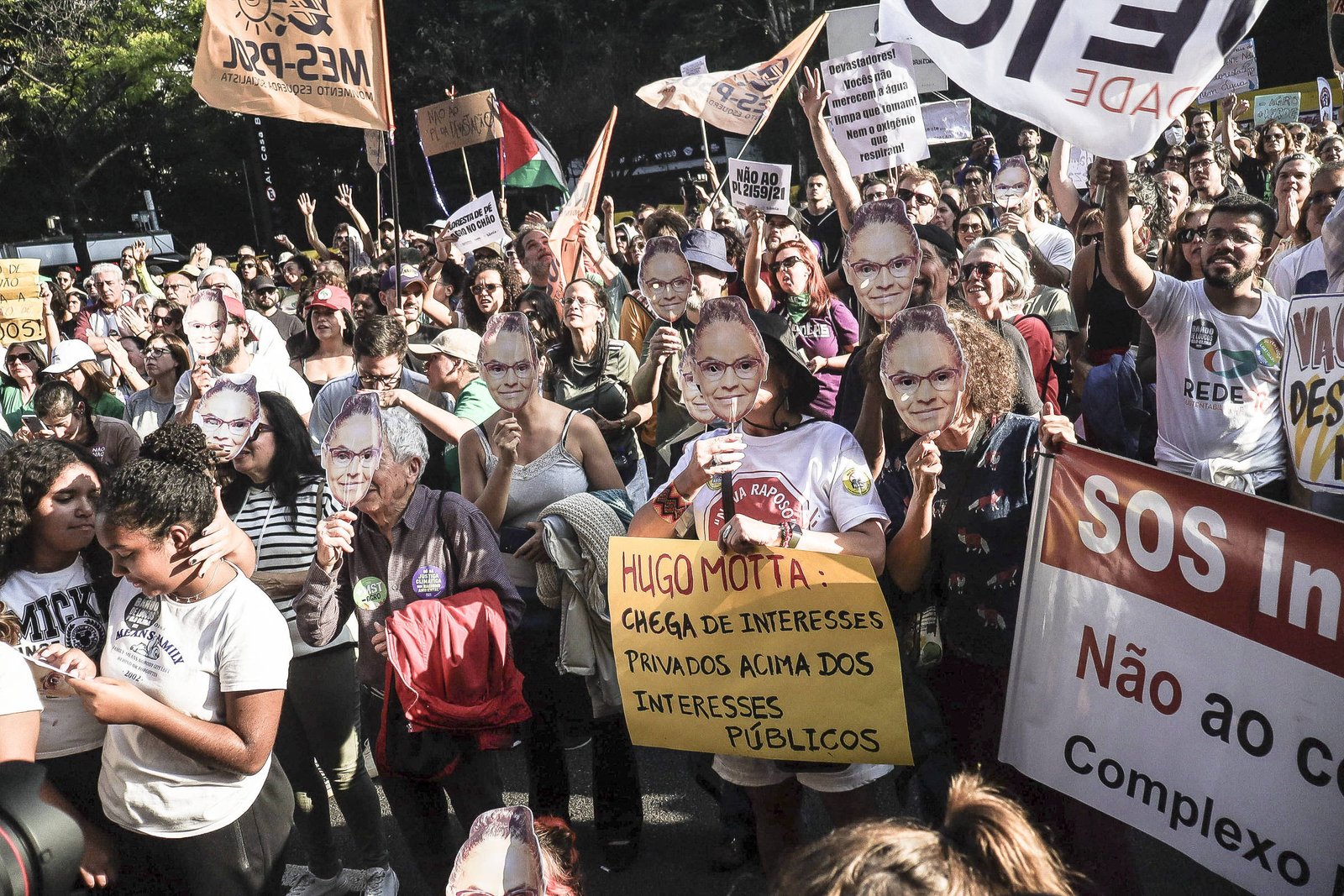 A imagem mostra uma grande manifestação com várias pessoas segurando cartazes e faixas. Muitas delas usam máscaras com o rosto de uma pessoa, possivelmente um político. Um dos cartazes destaca o nome