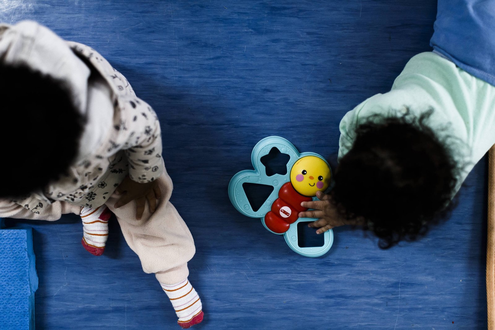 A imagem mostra duas crianças pequenas brincando em um tapete azul. Uma criança está de costas, vestindo uma blusa com estampa e calças claras, enquanto a outra criança está agachada, segurando um brinquedo colorido em forma de flor com uma figura sorridente no centro. Ambas as crianças têm cabelo cacheado e estão concentradas na brincadeira. A foto é feita de cima para baixo, então não é possível identificá-las.