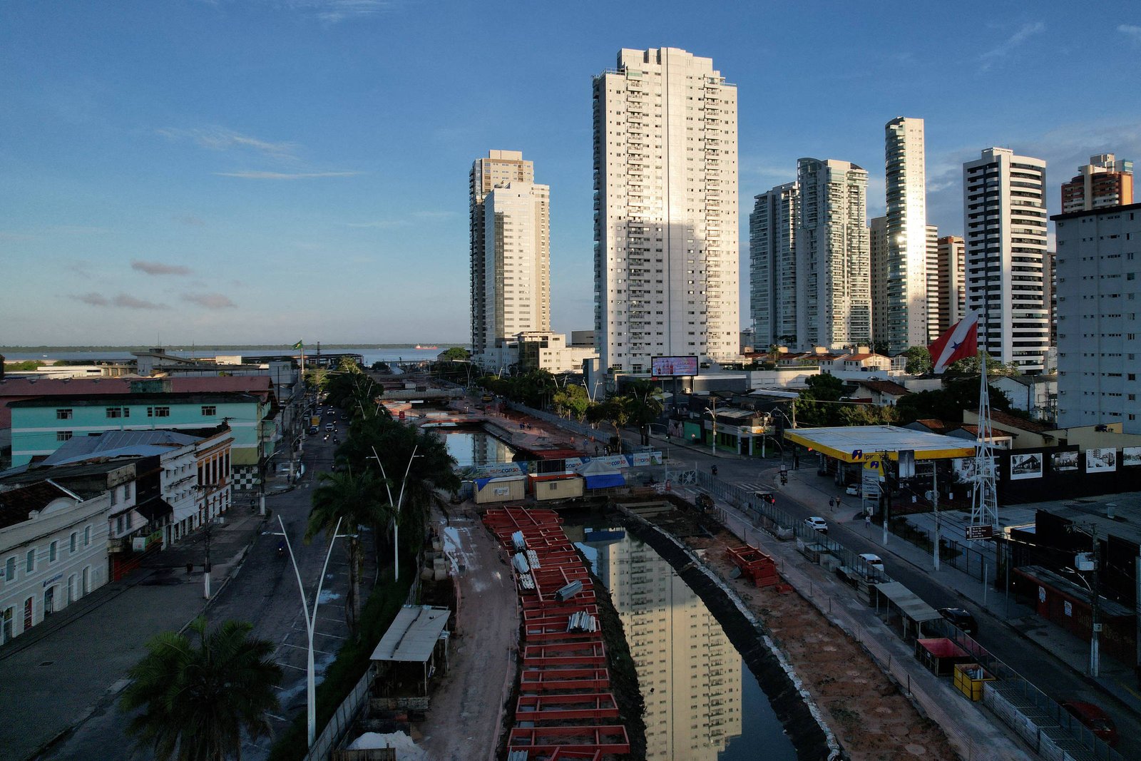 A imagem mostra uma vista urbana com arranha-céus altos à direita, refletindo a luz do sol. À esquerda, há edifícios de menor altura e uma área com vegetação. No centro, um canal é visível, com barcos ancorados e uma estrada ao lado. O céu está claro, indicando um dia ensolarado.