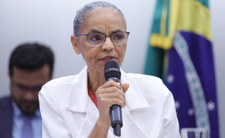 Ministra do Meio Ambiente, Marina Silva, fala ao microfone. Ela é uma mulher mulata, tem cabelos grisalhos presos num coque, usa óculos e veste um blazer branco. Ao fundo, do lado direito, há uma bandeira do Brasil