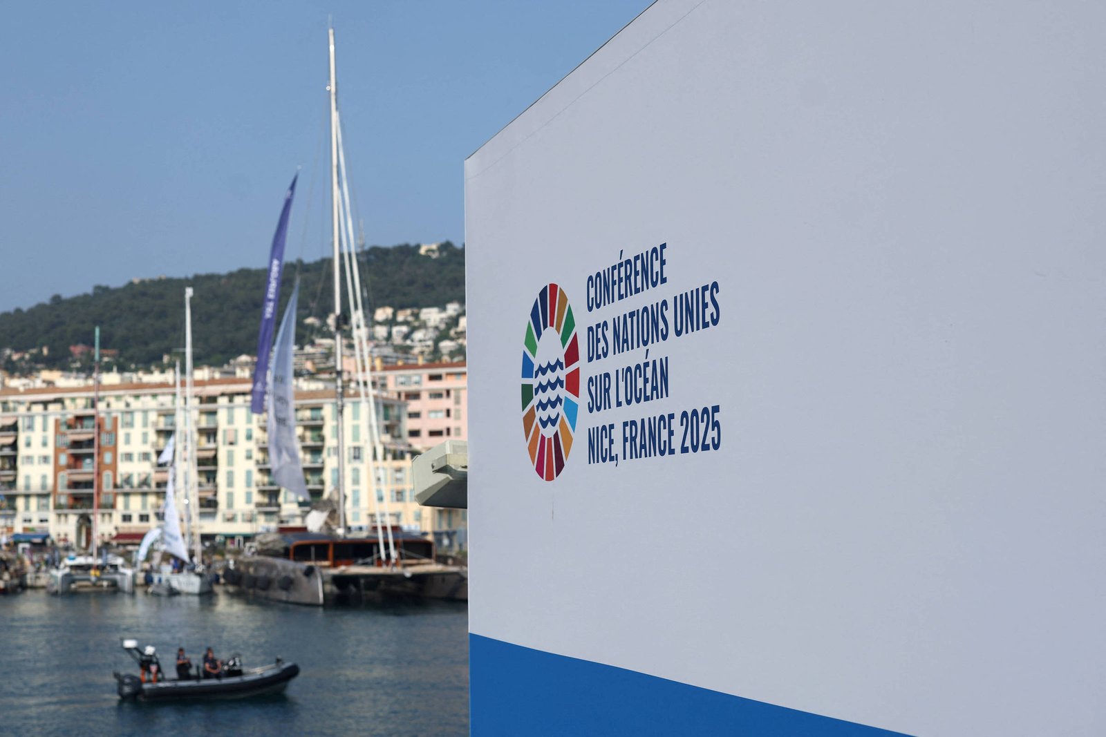 A imagem mostra um painel com o logotipo da Conferência das Nações Unidas sobre o Oceano, que ocorrerá em Nice, França, em 2025. Ao fundo, é possível ver um porto com barcos e edifícios à beira-mar, além de montanhas ao longe. O céu está claro e a cena transmite uma atmosfera de evento ao ar livre.