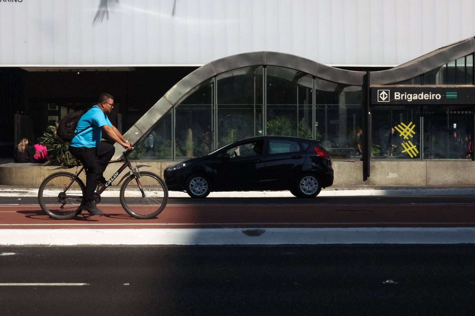 A imagem mostra um ciclista pedalando em uma ciclovia, ao lado de uma rua onde um carro preto está estacionado. Ao fundo, há uma estrutura arquitetônica com um design curvo e moderno, e uma estação de metrô com o nome