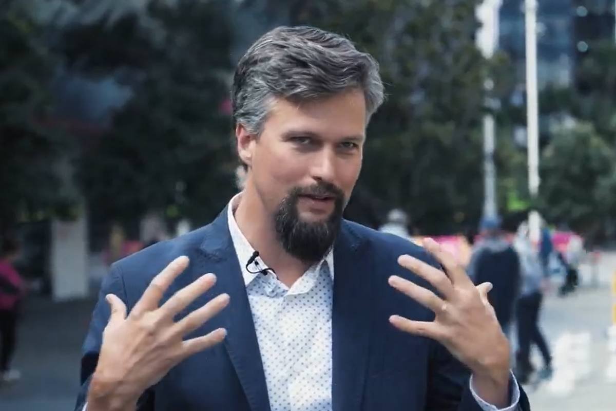 Um homem com cabelo grisalho e barba está em um ambiente urbano, gesticulando com as mãos enquanto fala. Ele usa um paletó escuro e uma camisa clara com padrões. Ao fundo, há pessoas caminhando e algumas árvores, além de estruturas urbanas visíveis.