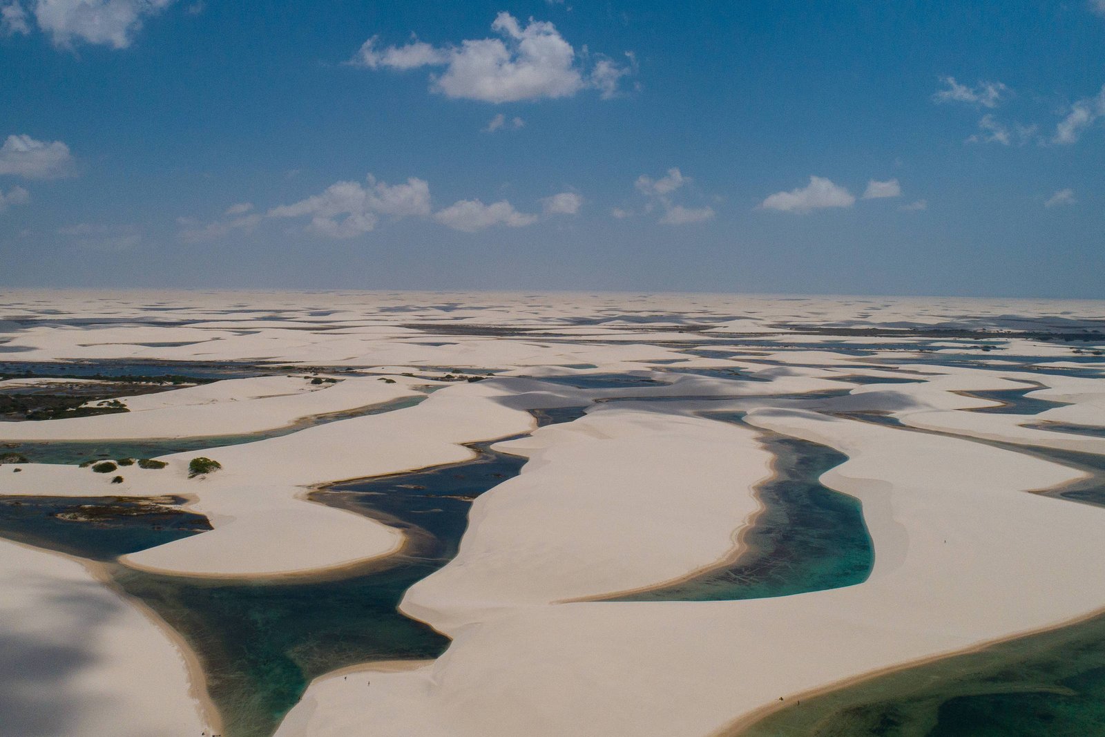 A imagem mostra uma vasta paisagem dos Lençóis Maranhenses, com dunas de areia branca e lagoas de água cristalina. O céu está claro, com algumas nuvens dispersas, e a areia se estende em padrões ondulados, intercalados por áreas de água. A vegetação é escassa, visível em algumas partes da imagem.