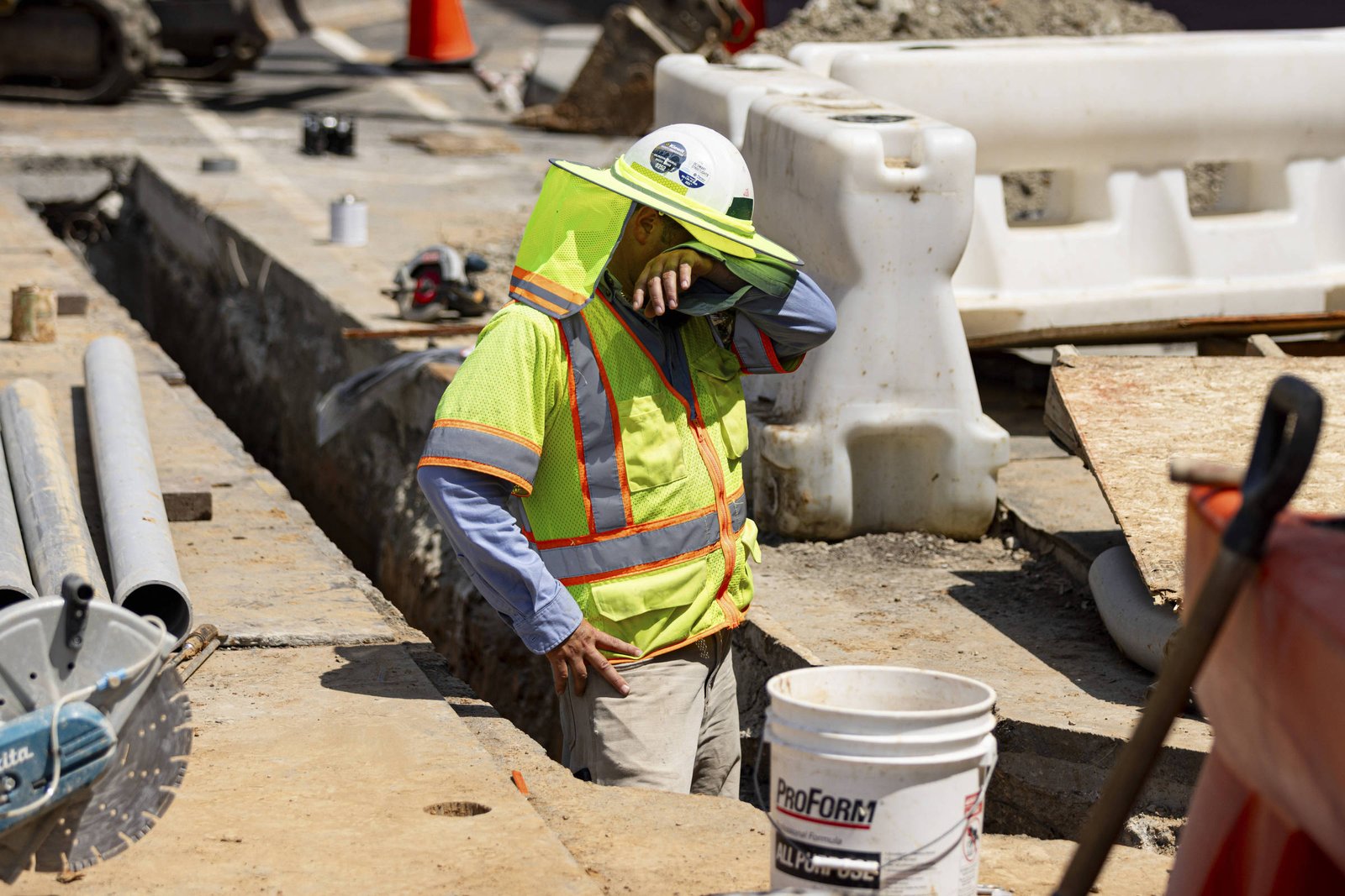 Um trabalhador de construção civil está em um canteiro de obras, usando um colete de segurança amarelo e um chapéu. Ele enxuga o suor do rosto com a mão. Ao fundo, há uma vala aberta, equipamentos de construção e barreiras de segurança. O ambiente é ensolarado e há materiais de construção ao redor, como tubos e baldes.