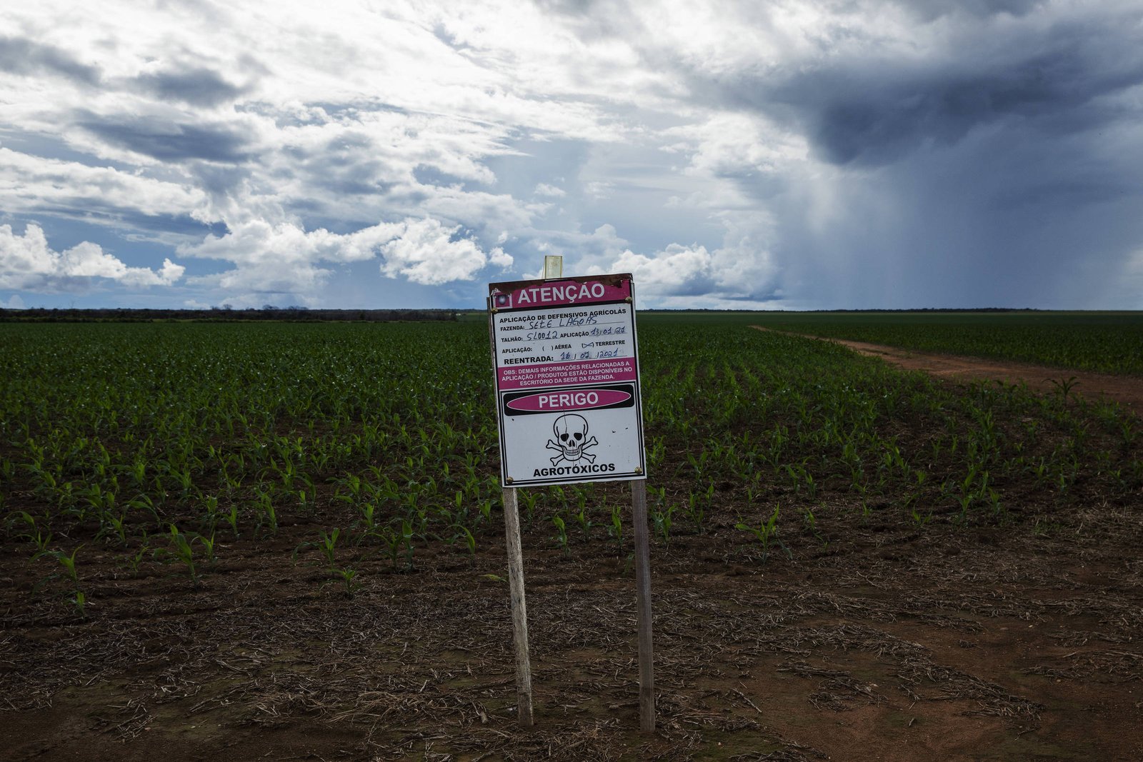 A imagem mostra uma placa de aviso em um campo verde. A placa está posicionada em um terreno de terra batida e apresenta um fundo de céu nublado com nuvens escuras. A placa contém informações de advertência, mas o texto não é legível na imagem.