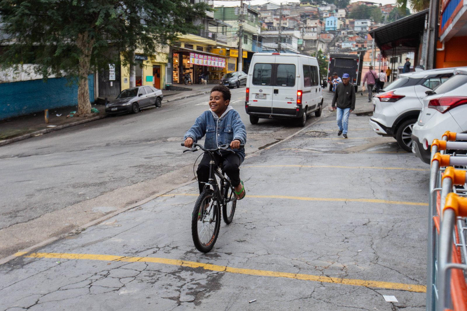 Uma criança está andando de bicicleta em uma rua urbana. O menino sorri enquanto pedala. Ao fundo, há um furgão branco estacionado e algumas pessoas caminhando. O cenário é de uma área com casas e comércio.