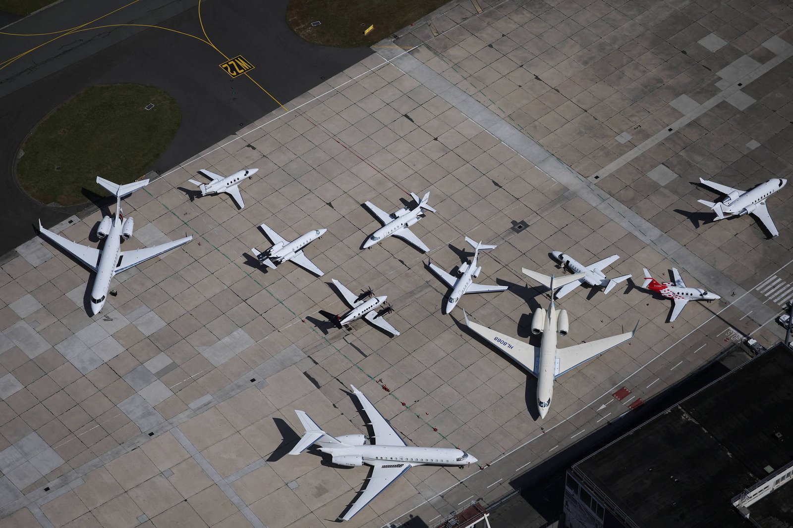 Uma visão aérea de um pátio de aeroporto com várias aeronaves estacionadas. Há diferentes tipos de jatos, todos predominantemente brancos, dispostos em várias posições. O chão é de concreto, com marcas de pista visíveis. Ao fundo, há uma área verde e marcas de taxiamento.