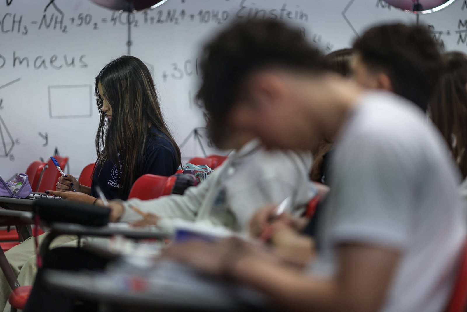 A imagem mostra um grupo de estudantes em uma sala de aula. No primeiro plano, uma estudante com cabelo longo e escuro está concentrada, escrevendo em um caderno. Ao fundo, outros estudantes estão visíveis, alguns com a cabeça baixa, também escrevendo. As mesas são de cor escura e as cadeiras são vermelhas. As paredes da sala têm anotações e fórmulas científicas escritas, sugerindo um ambiente acadêmico.
